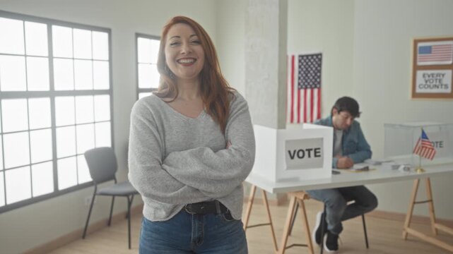 Woman arms crossed smiling by vote box with man at registration table and american flags in building; civic duty confidence.