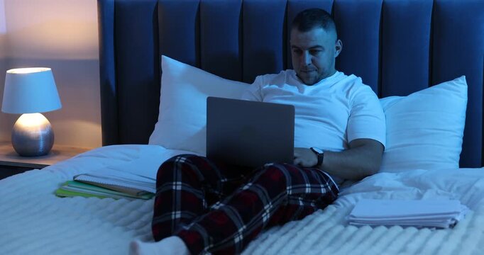 Workaholic. Overwhelmed man in pajamas working with laptop between stacks of documents on bed indoors at night. Camera moving forward