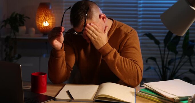Workaholic. Overwhelmed man at wooden desk with laptop and notebook indoors at night, camera moving forward