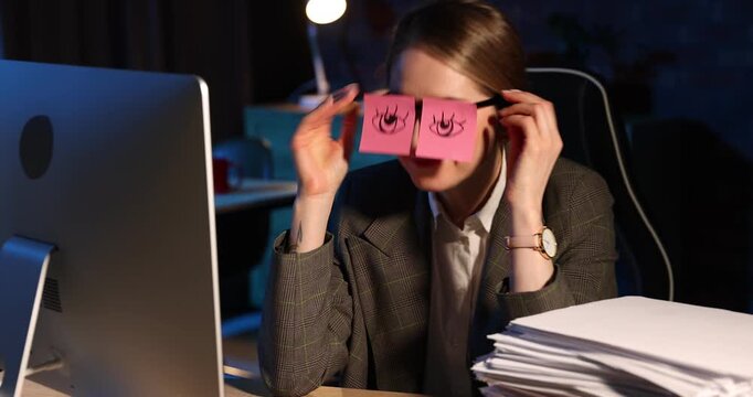 Workaholic. Woman yawning and putting on glasses with fake eyes drawn on sticky notes near stack of documents and computer at wooden desk indoors at night. Camera moving forward