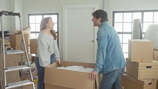 Couple lifting a large cardboard box filled with packing peanuts, woman tosses foam peanuts toward laughing man in building; playful togetherness.