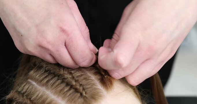 Hairstylist braiding teenage girl's hair indoors, closeup