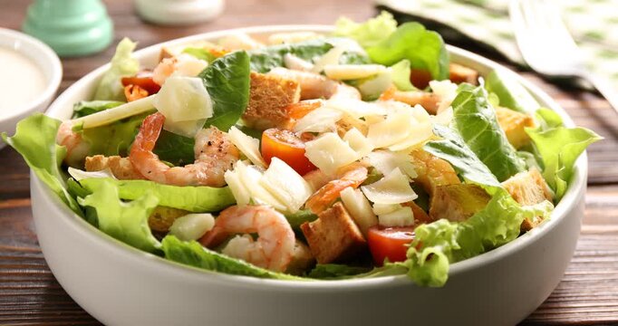 Woman adding cheese into bowl of delicious Caesar salad with shrimps at wooden table, closeup