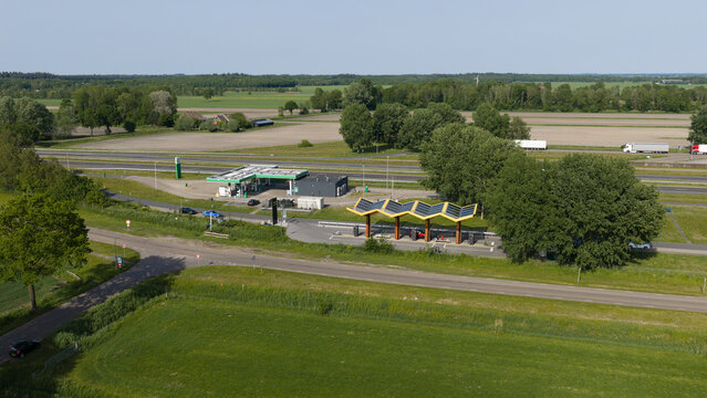 Darp, Netherlands - 23 May 2026: Aerial view of Fastned Charging Station with its yellow solar roof and a nearby gas station along a highway surrounded by green fields and trees.