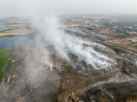 Aminbazar, Bangladesh - 21 February 2026: Aerial view of the Aminbazar landfill with thick white smoke rising from burning waste, excavators working on the soil, and birds circling.