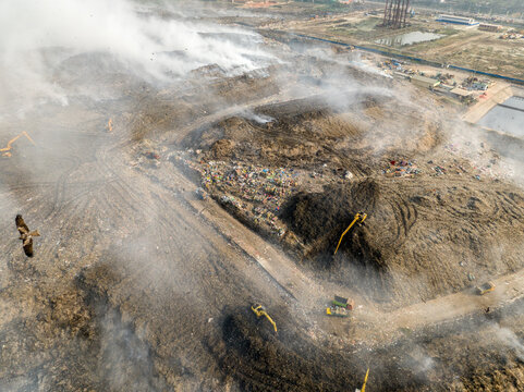 Aminbazar, Bangladesh - 21 February 2026: Aerial view of the Aminbazar landfill with smoke rising from waste piles, excavators operating on site, and a bird flying in the foreground.