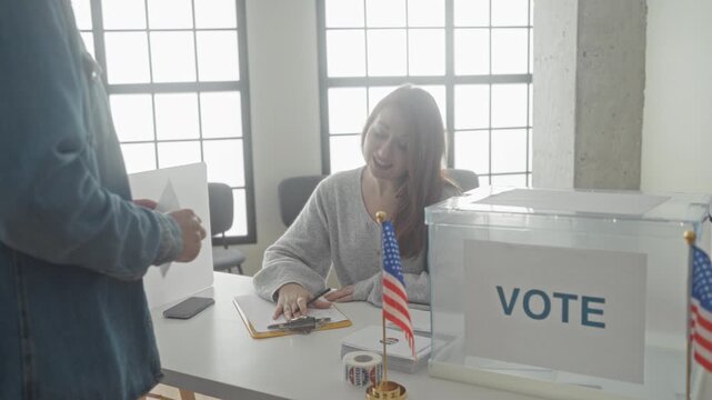 Woman placing a ballot into a clear ballot box at a pollingstation, reaching across a table with clipboard and american flags visible; responsibility participation.