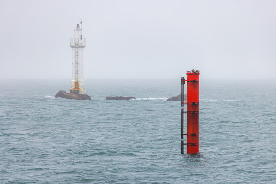 View of a white lighthouse on a rocky outcrop and a red beacon in the choppy sea under a foggy sky in Ushant, Brittany, France.