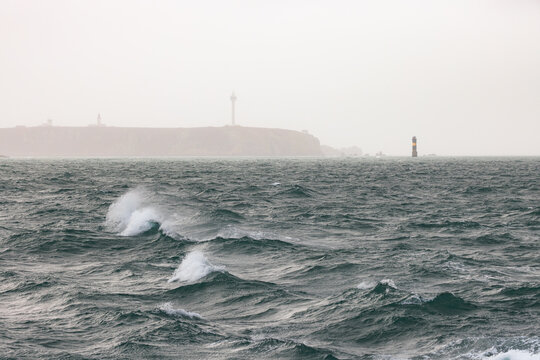 View of rough sea waves with whitecaps, a distant lighthouse on the misty cliffs and a beacon tower in the water at Ushant, Brittany, France.