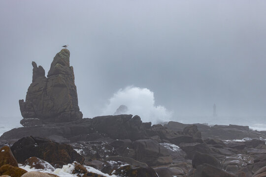 View of powerful waves crashing against a rocky coastline with a jagged rock formation and a distant lighthouse in the mist in Ushant, Brittany, France.