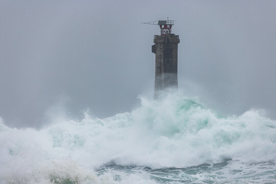 View of a tall stone lighthouse standing amidst powerful crashing waves and sea spray under a stormy grey sky in Ushant, Brittany, France.
