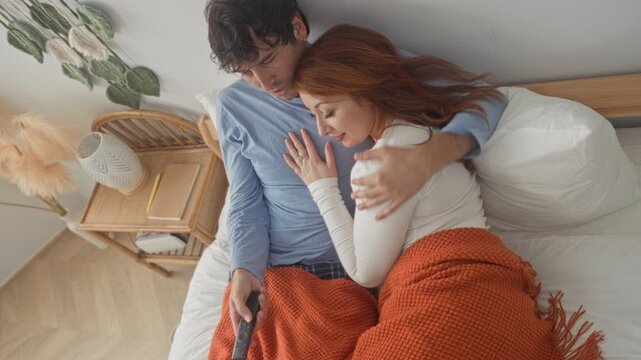 Man holding tv remote while embracing woman on bed in a bedroom inside a building, watching together under an orange knit blanket; comfort connection.