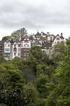 View of the historic Ramsay Garden buildings with white facades and red timber framing nestled behind lush green trees under an overcast sky in Edinburgh, Scotland, United Kingdom.