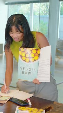 Vertical video: Opening cookbook, woman in lime-green top presenting recipes and noting in notebook