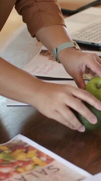 Vertical video: Reaching woman placing apple, avocado, brochures making layout at table tan sleeves