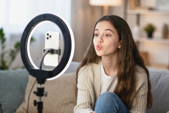 A young teenage female creator sits on a home sofa, using a ring light and smartphone to record live streaming or vlog content with a pouting expression.