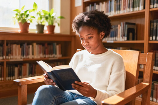 A focused Black teenage girl sits in a wooden armchair reading a hardcover book inside a sunlit school library.