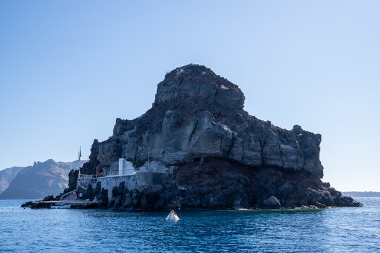 View of the Skaros Rock volcanic formation with a small white structure at its base under a clear blue sky in Fira, Greece.