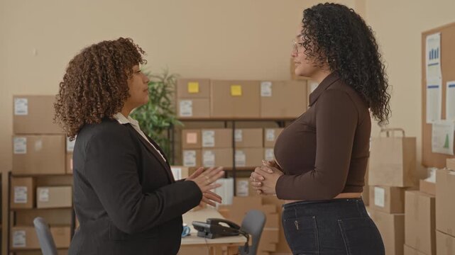 Two women small business workers clasp hands and stand with arms crossed amid stacked shipping boxes and parcels in a storage room inside a building; confidence teamwork.