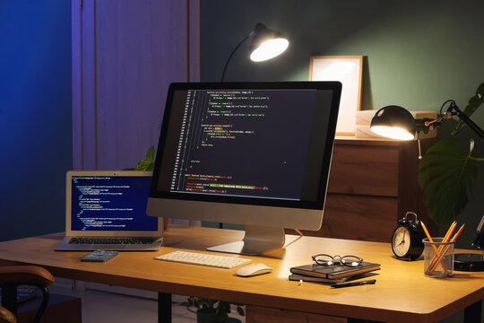 Programmer's workplace with computer, laptop and glowing lamps in dark office