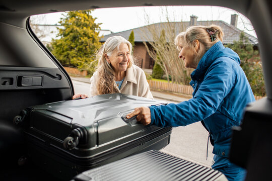 Smiling senior and woman loading suitcase into car trunk on suburban street