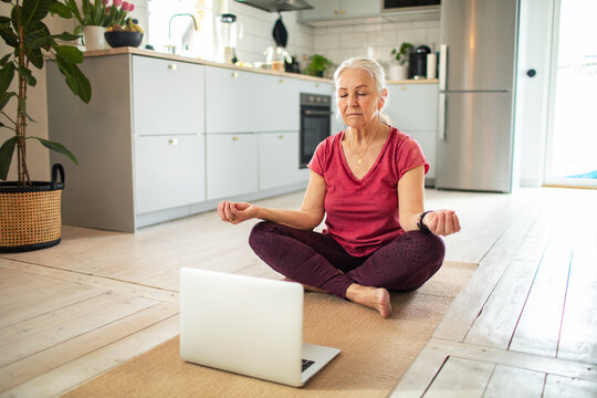 Senior woman meditating with laptop in home kitchen