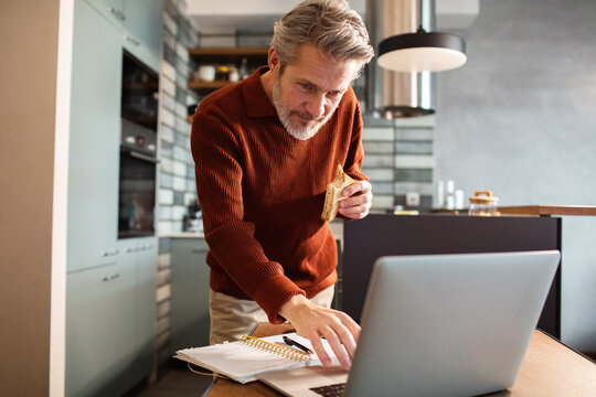 Mature man working on laptop while eating sandwich in home kitchen