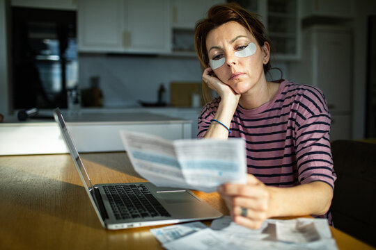 Tired woman with eye patches reviewing bills in home kitchen