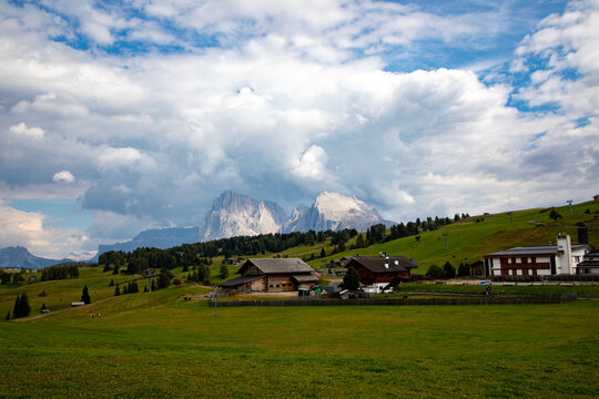 View of traditional wooden chalets in a lush green alpine meadow with the jagged peaks of the Dolomites under a cloudy sky in Rocca Pietore, Veneto, Italy.