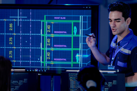 A construction engineer uses a tablet to review a BIM (Building Information Modeling) file. A CAD blueprint of a residential smart building is on a screen in the control center.