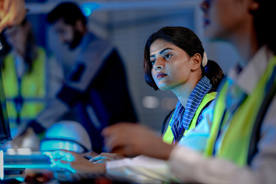 An Indian female technician in a safety vest monitors an AI-driven automation system. She is working in a smart factory command center, analyzing system data at night.