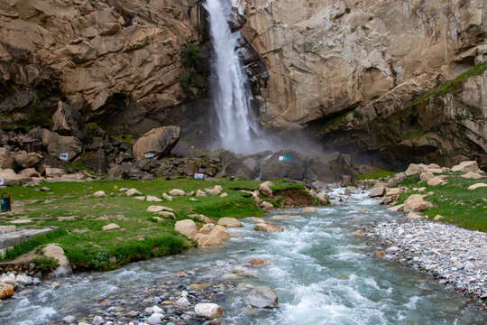 View of Khamosh Waterfall cascading down steep rocky cliffs into a clear mountain stream surrounded by green grass and boulders in Skardu, Gilgit, Pakistan.
