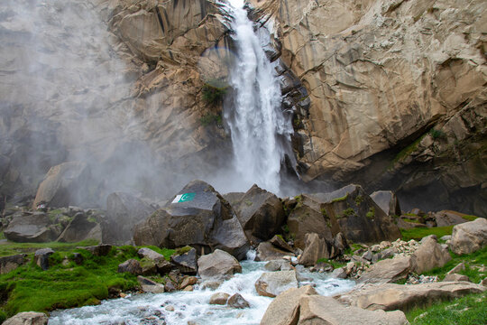 View of Khamosh Waterfall cascading down steep rocky cliffs into a flowing stream with large boulders and green grass in Skardu, Gilgit, Pakistan.