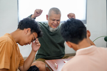 Fototapeta premium Cheerful Senior Asian Man Raising His Fist in Celebration While Winning Domino Game With Young Men