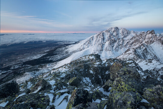 View of the snow-covered peaks of Slavkovsky stit with a rocky foreground and a sea of clouds under a soft sunset sky in Vysoke Tatry, Presovsky kraj, Slovakia.