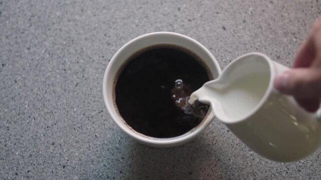 The process of pouring milk or white cream from a small ceramic container into a cup of strong black coffee.