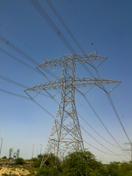 View of a high-voltage electricity pylon and power lines against a clear blue sky with green bushes at the base UAE.