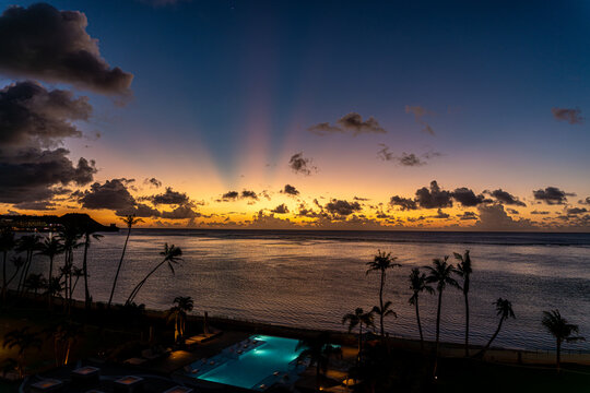 View of the sunset over Tumon Bay with palm trees, a lit swimming pool, and dramatic crepuscular rays in the sky Tumon, Guam, Guam.