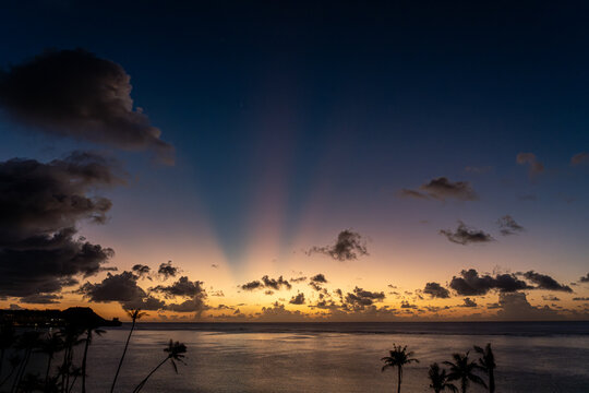 View of Tumon Bay at sunset with crepuscular rays stretching across the sky above palm tree silhouettes and calm ocean waters in Tumon, Guam, Guam.