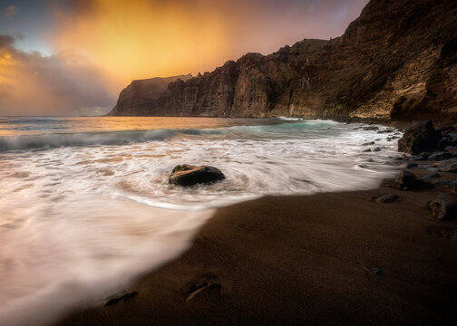 View of the dramatic cliffs of Los Gigantes at sunset with waves crashing on a black sand beach under a golden sky in Acantilados de Los Gigantes, Canarias, Spain.