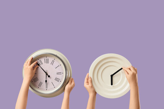 Female hands holding different wall clocks on lilac background
