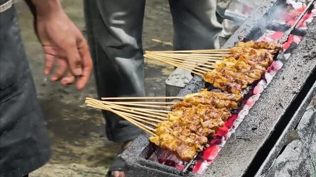 A dynamic and appetizing street food photograph showing two rows of traditional Indonesian satay skewers being grilled
