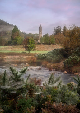 View of the Glendalough Round Tower standing amidst autumn trees and misty hills with a flowing river and spider webs in the foreground Glendalough, County Wicklow, Ireland.
