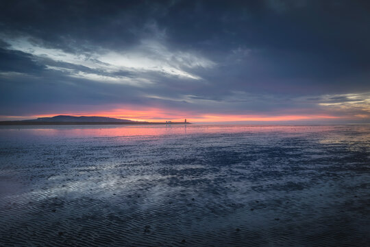 View of the sunset over the sea with the South Wall lighthouse in the distance under a dramatic cloudy sky County Dublin, Ireland.