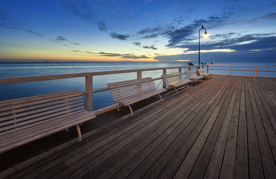 View of Orlowo Pier with white wooden benches and a glowing street lamp under a deep blue twilight sky over the Baltic Sea in Gdynia, Wojewodztwo pomorskie, Poland.