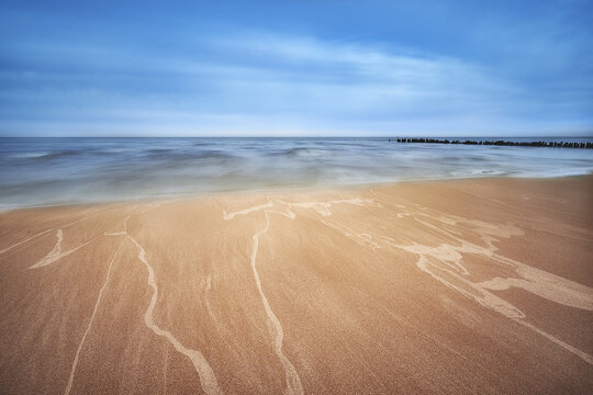 View of a sandy beach with sea foam patterns, gentle waves of the Baltic Sea, and a wooden breakwater under a blue sky in Debki, Pomeranian Voivodeship, Poland.