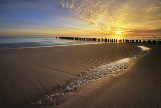 View of a golden sunset over the sandy shore with wooden breakwaters and a small stream on the beach in Chalupy, Wojewodztwo pomorskie, Poland.