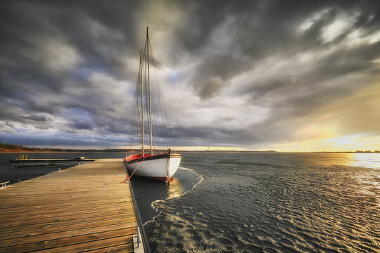 View of a sailboat moored at a wooden pier on the partially frozen Lake Tarnobrzeg under a dramatic sunset sky in Tarnobrzeg, Podkarpackie, Poland.