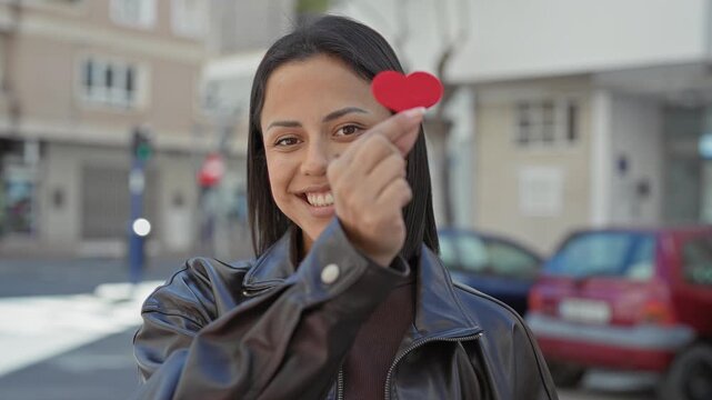 Woman holding a small red paper heart between her fingers, smiling directly to camera on a city street with parked cars and buildings; affection connection.