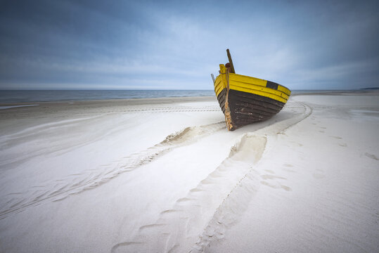 View of a traditional yellow and brown wooden fishing boat resting on a sandy beach with tire tracks under a cloudy sky in Debki, Pomeranian Voivodeship, Poland.
